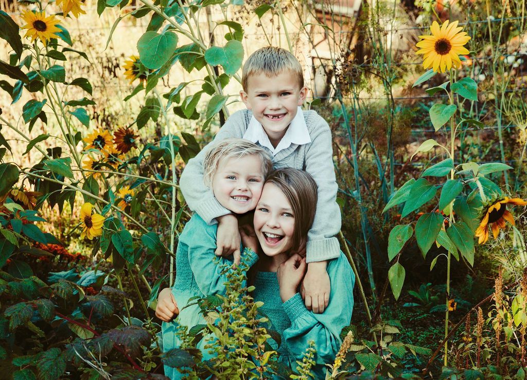 Three smiling children stacked in a garden of sunflowers.