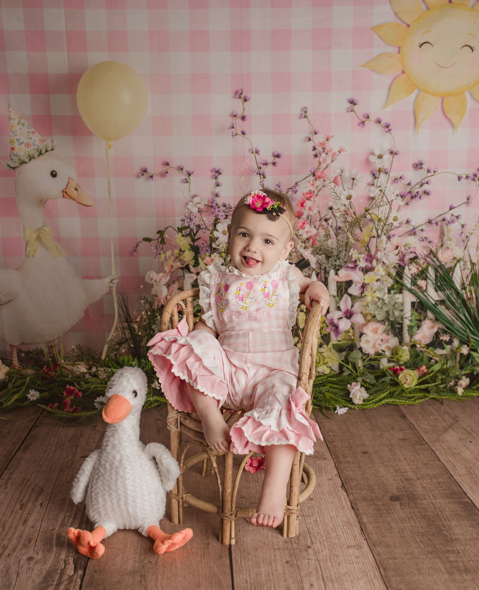 Child in pink dress sitting on a chair with a stuffed goose, in a floral spring-themed backdrop.