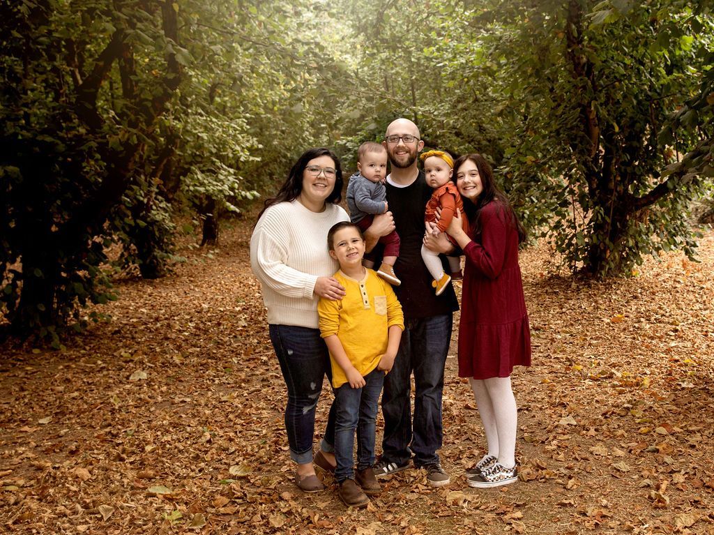 Family of six posing in a forest of fall leaves. Two adults hold two young children; older children stand on either side.