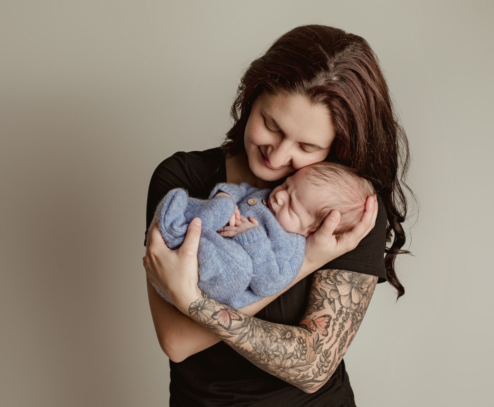 Woman with arm tattoos cradles a swaddled baby, smiling. Neutral background.