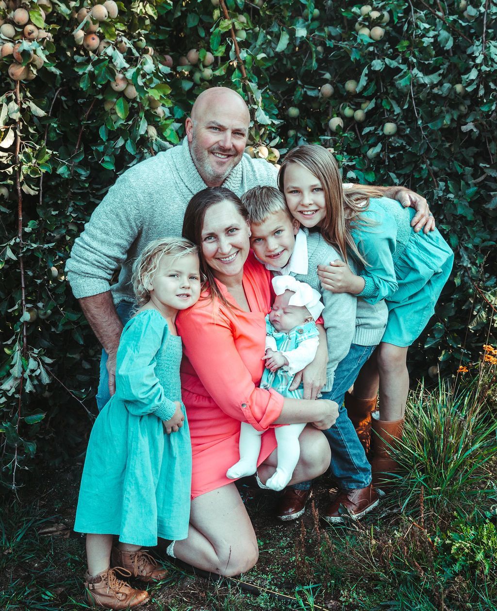 Family of six in front of apple tree; smiling. Mother in orange, father in gray, children in blue and gray.