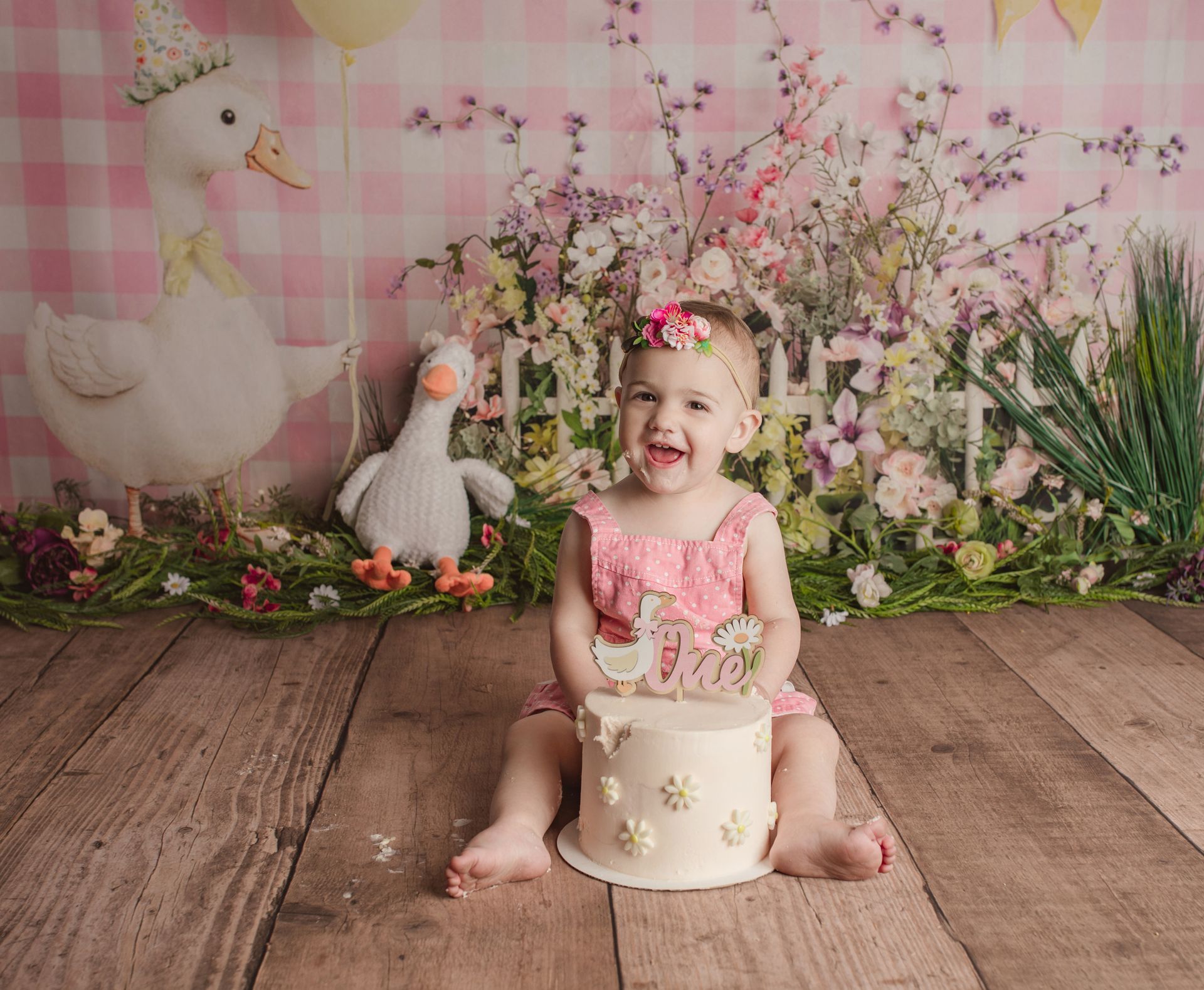 Smiling child in pink dress holding a cake, seated on wooden floor with spring floral backdrop and lamb decorations