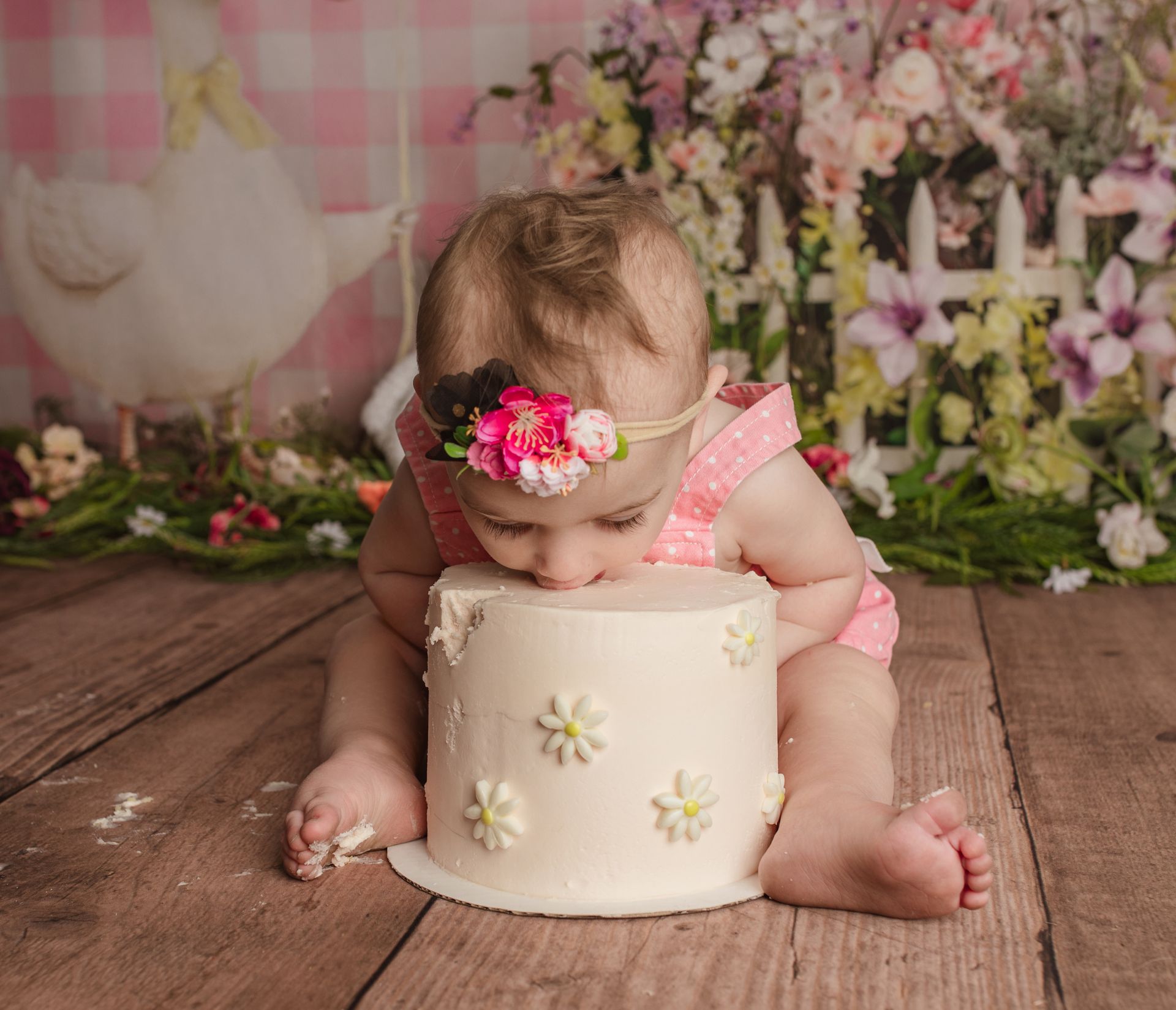Baby in pink outfit leaning over a white floral cake in a rustic, flower-filled backdrop