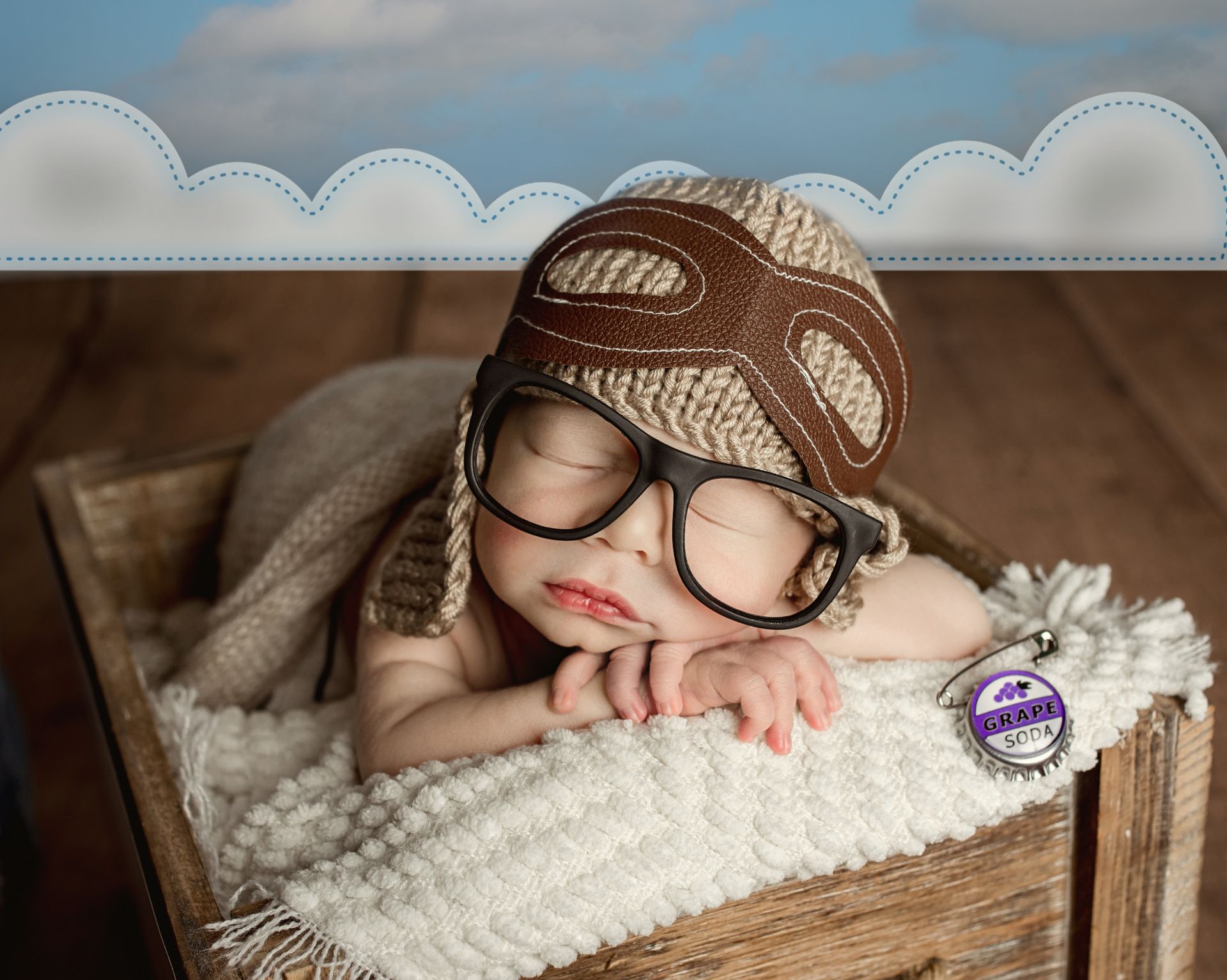 Newborn in wooden box, wearing aviator hat and glasses, sleeping peacefully.
