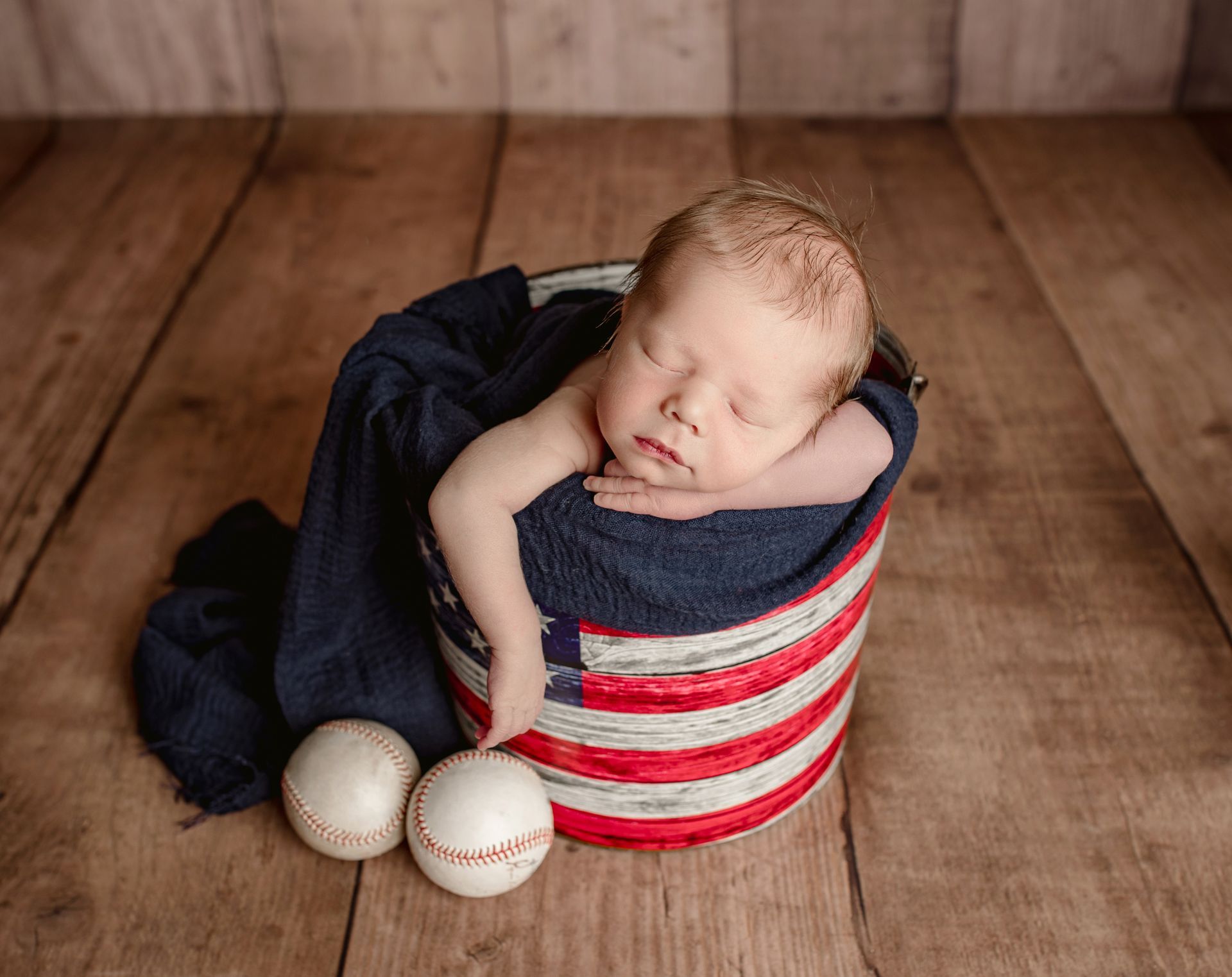 Baby asleep in American flag-decorated bucket, with two baseballs on wooden surface.