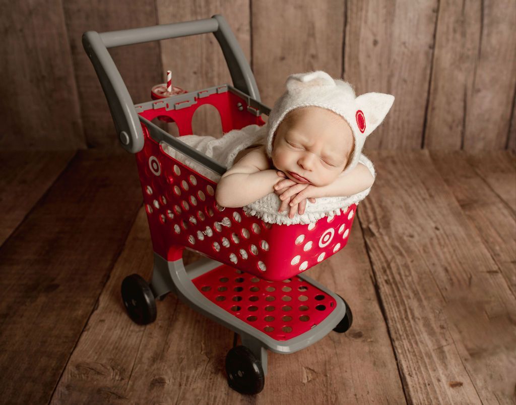 Newborn in a red toy shopping cart wearing a white cap, sleeping on a wood background.