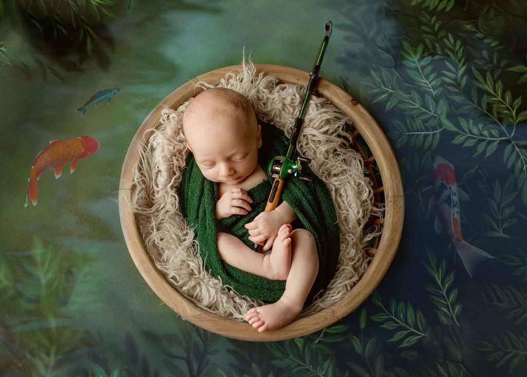 Newborn baby wrapped in green blanket, sleeping in a wooden bowl with fishing rod, koi fish in background.