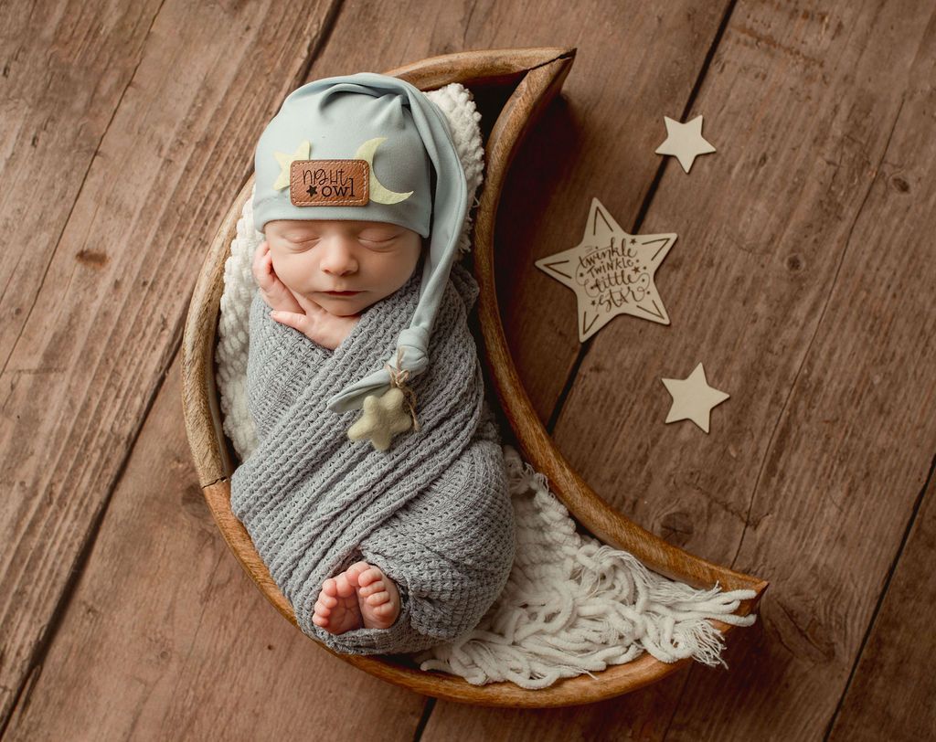 Newborn baby wrapped in gray, sleeping in wooden moon, wearing a blue hat, with star decorations.