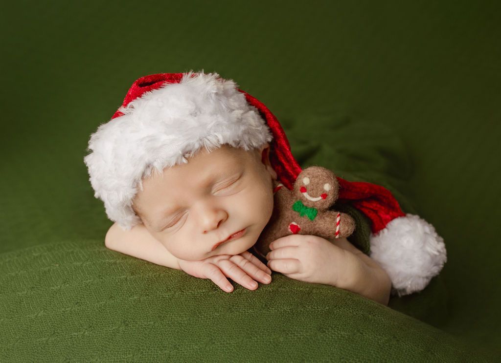 Newborn wearing Santa hat, sleeping on green blanket, clutching gingerbread man.