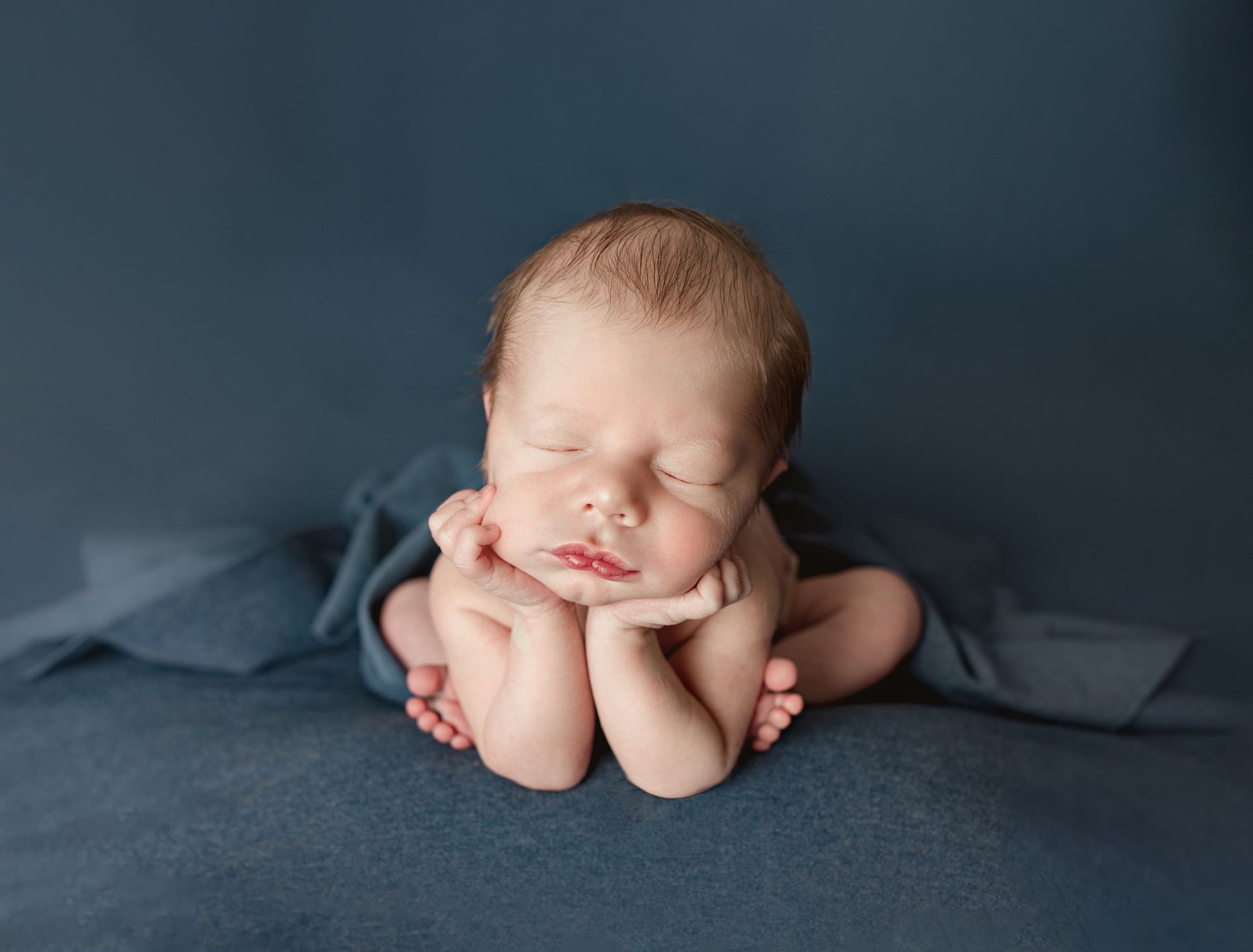 Newborn baby sleeping, head resting on hands, wrapped in blue fabric.