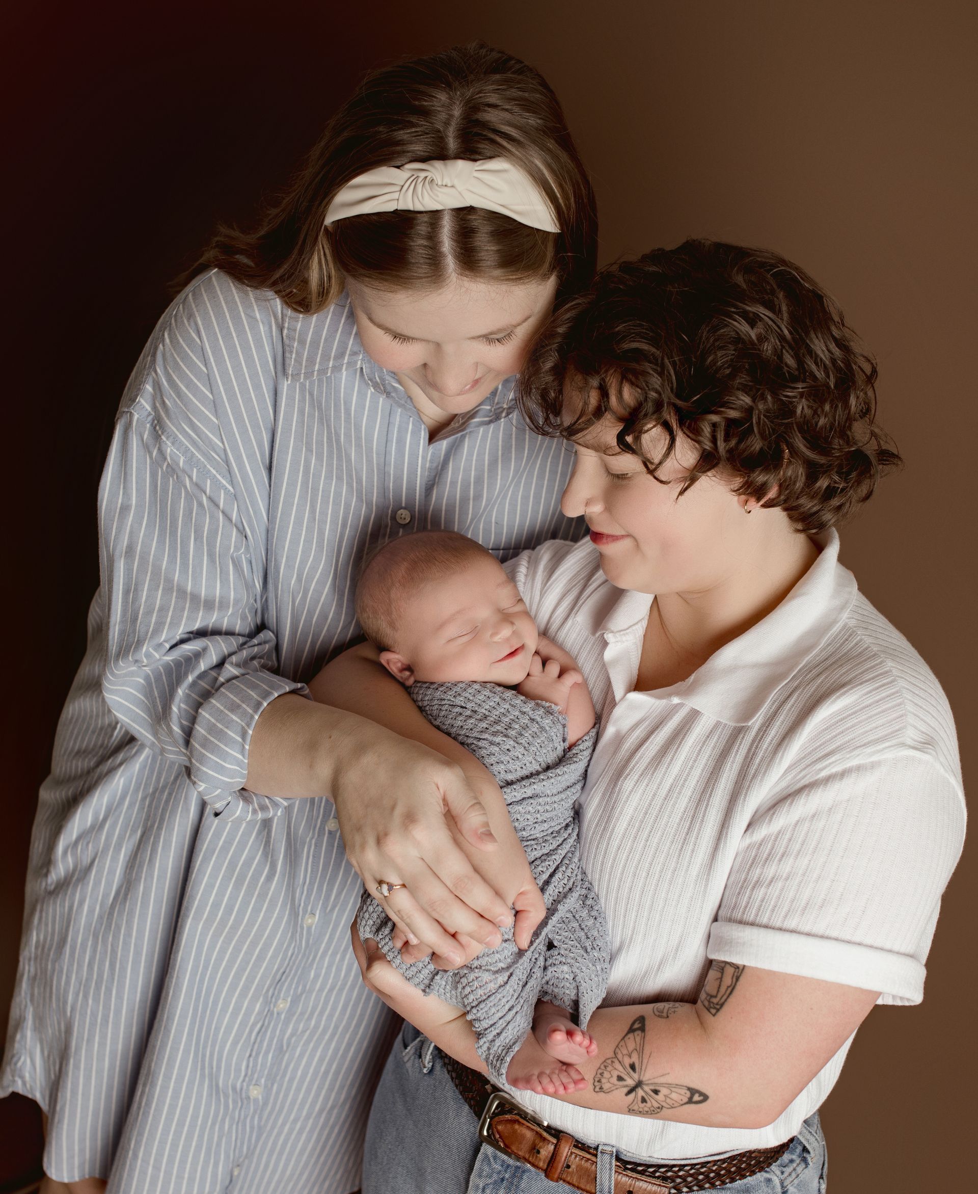 Two women cradle a swaddled infant against a brown backdrop.