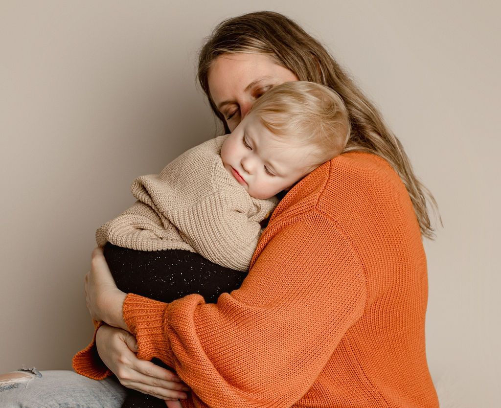 Woman in orange sweater holding a sleeping child wearing a tan sweater.