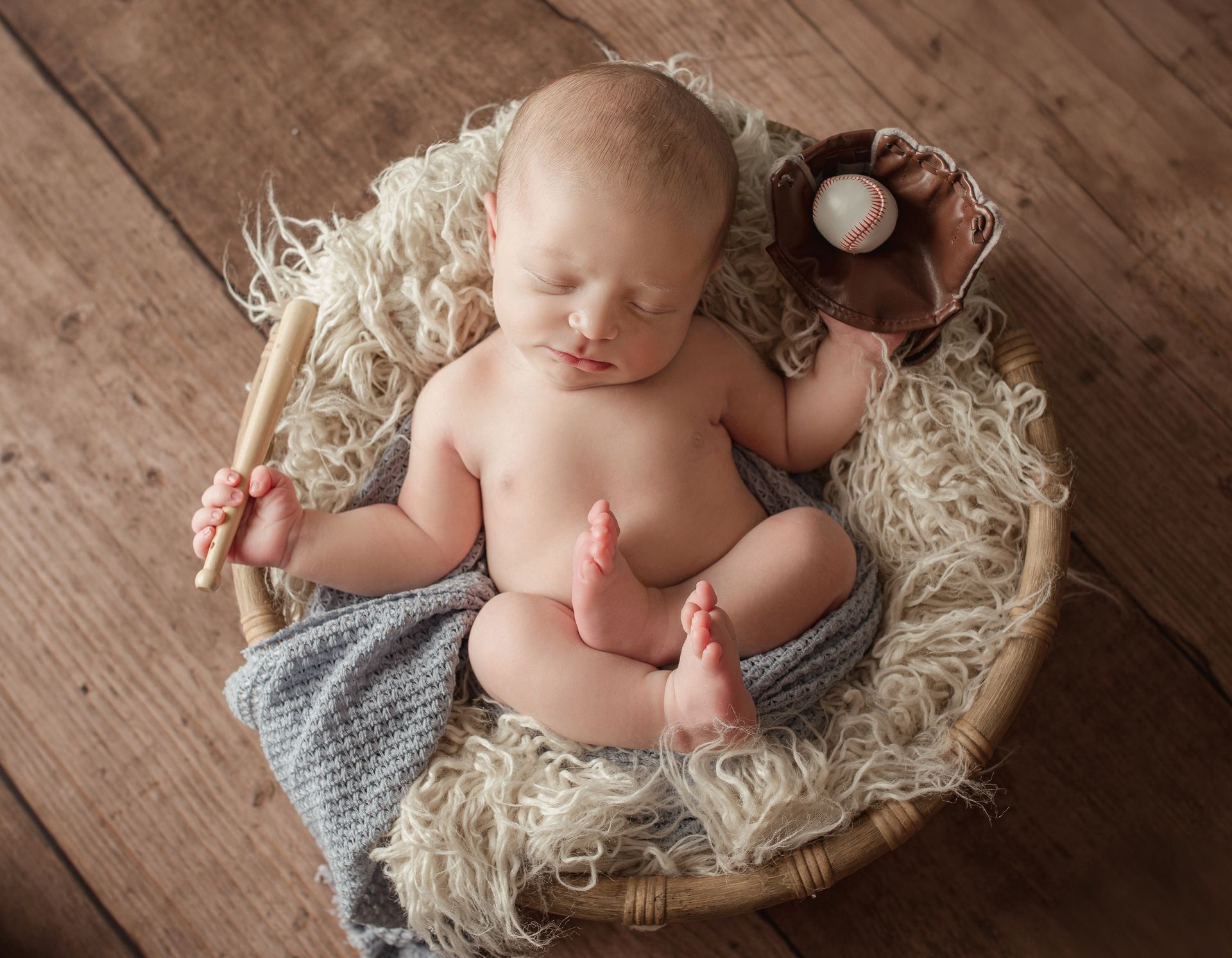 Newborn baby sleeping in a woven basket with baseball bat, glove, and ball. Wooden background.
