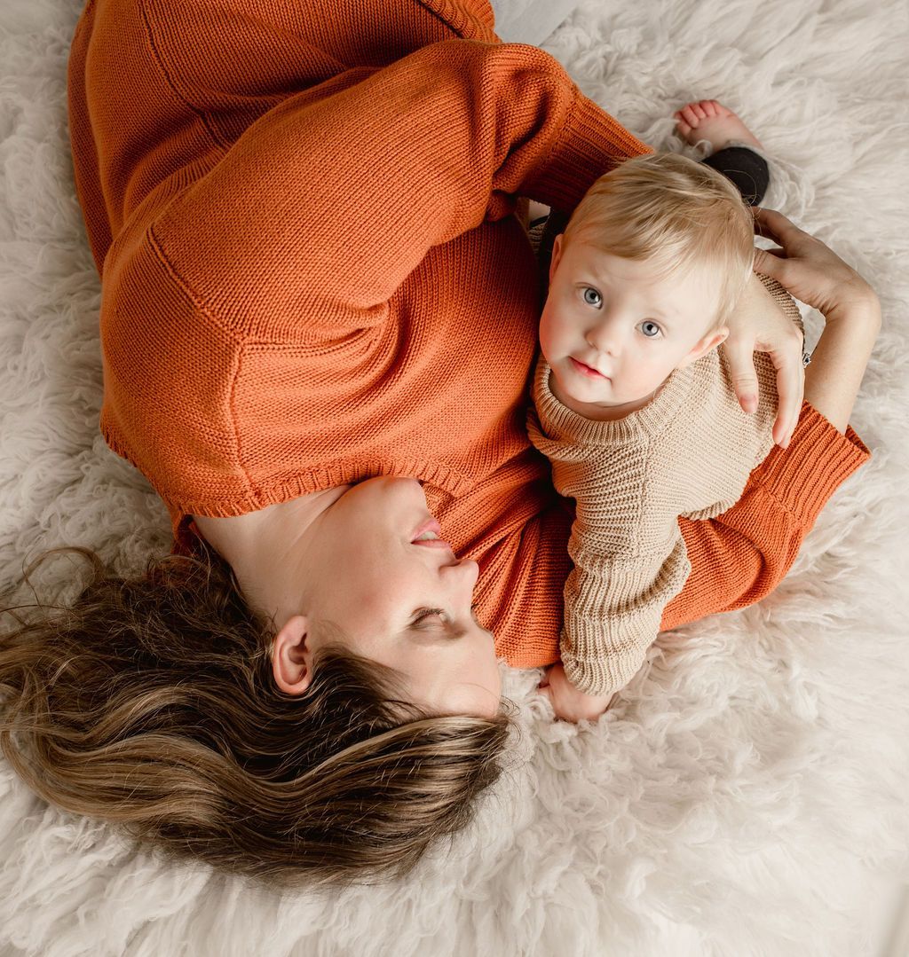 Woman and baby lying on fluffy white rug, both wearing knit sweaters, looking at camera.