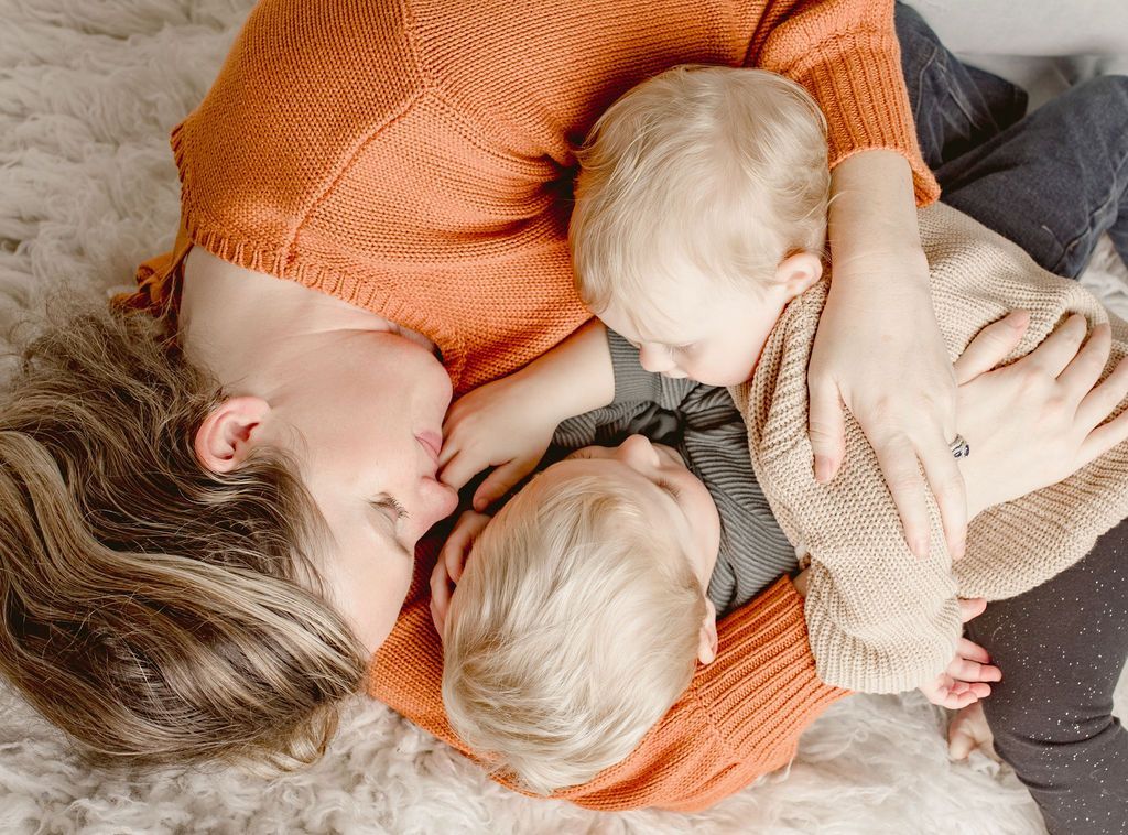 Woman in orange sweater embraces two toddlers on a fluffy rug.