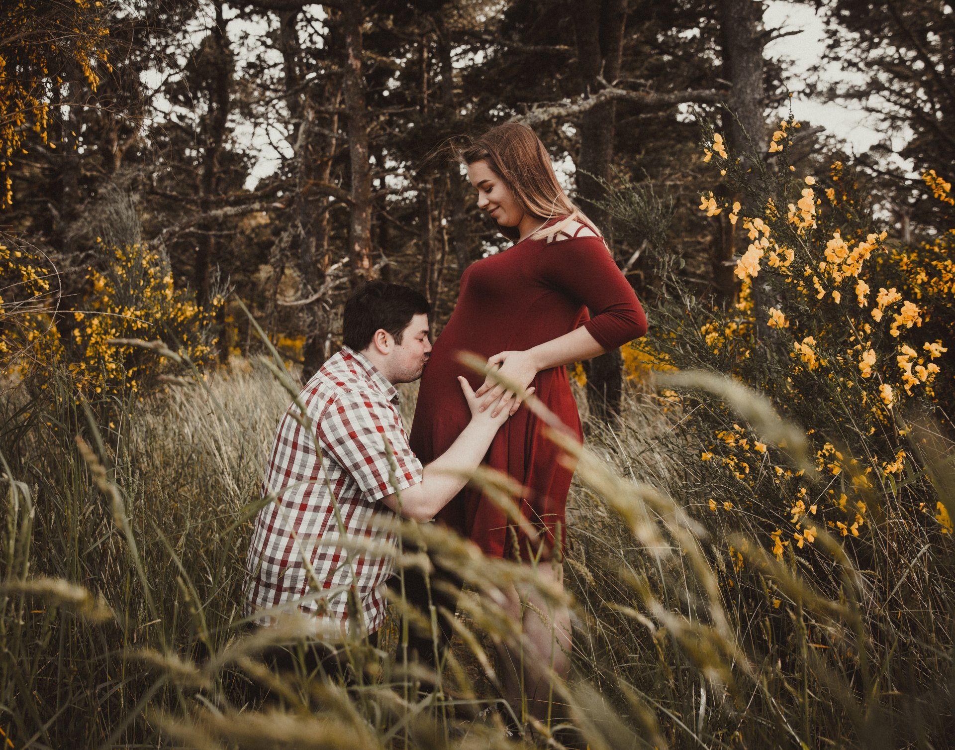 Man kneels, kissing pregnant woman's belly in tall grass. Woman in red dress smiles in wooded area.