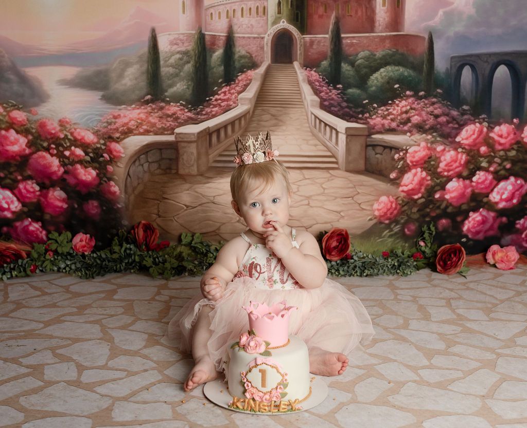 A baby with a crown eats cake in front of a painted castle and rose garden.