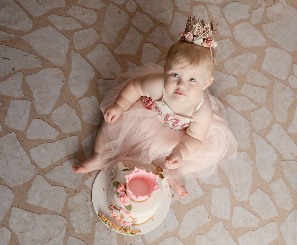 Baby in pink dress and crown sits beside a small cake on stone floor.