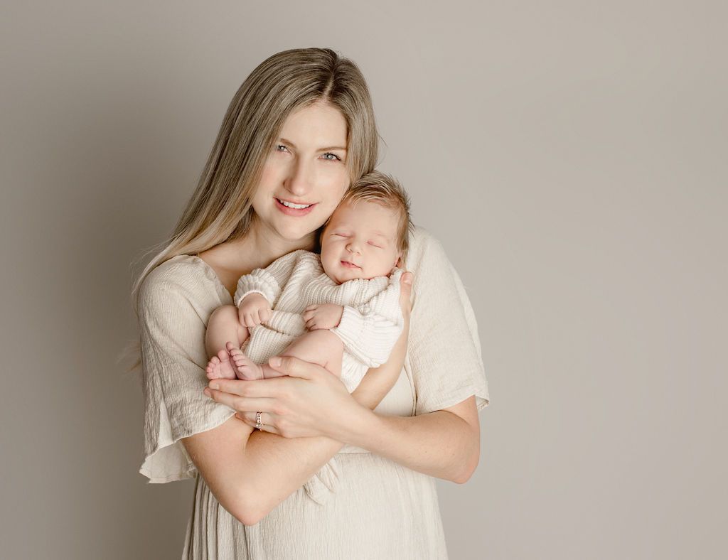 Woman holding a newborn baby, both wearing neutral-colored clothing, smiling in front of a gray backdrop.