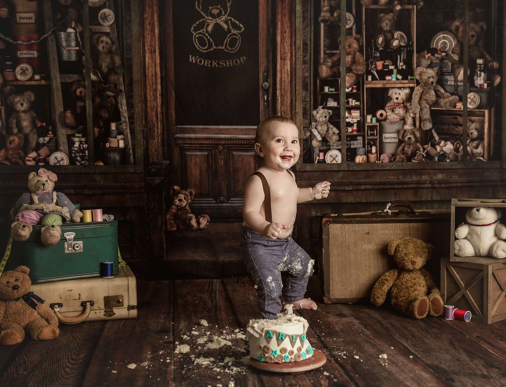 Smiling baby with cake smeared on clothes celebrates first birthday in front of teddy bears.