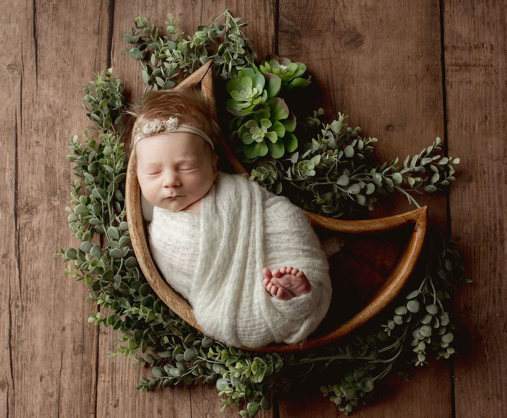 Newborn wrapped in a white blanket, in a wooden bowl with greenery, on a wooden surface.
