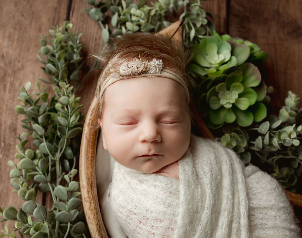 Newborn swaddled in cream blanket, headband with small bow, surrounded by greenery in a wooden bowl.