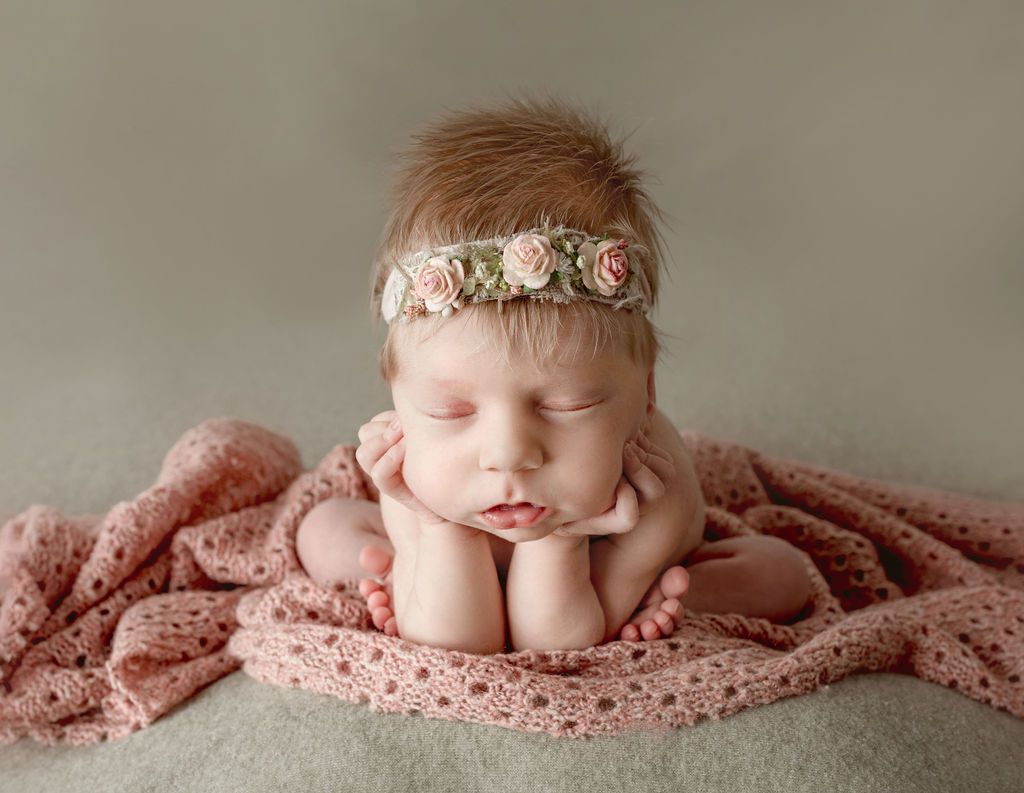 Newborn with closed eyes, hands under chin, wearing floral headband, on pink blanket.