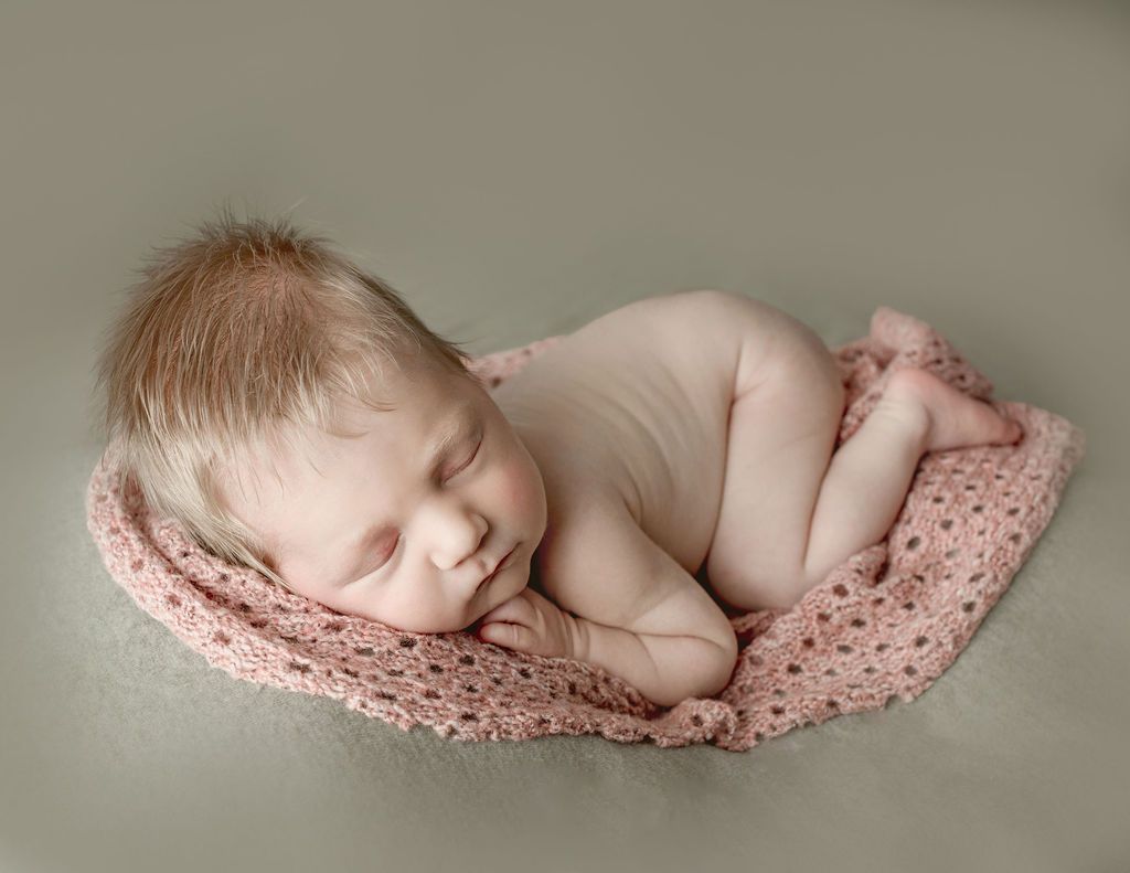 Newborn baby sleeping curled up on pink crocheted blanket.