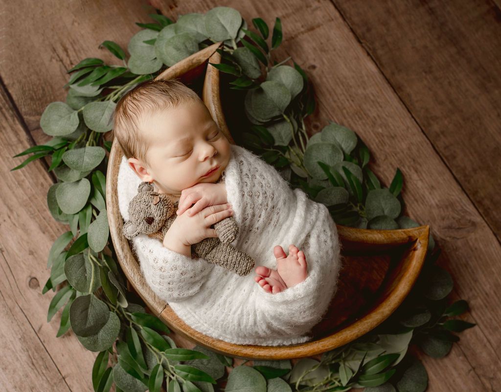 Newborn wrapped in a white blanket, cuddling a teddy bear, nestled in a wooden moon surrounded by greenery.
