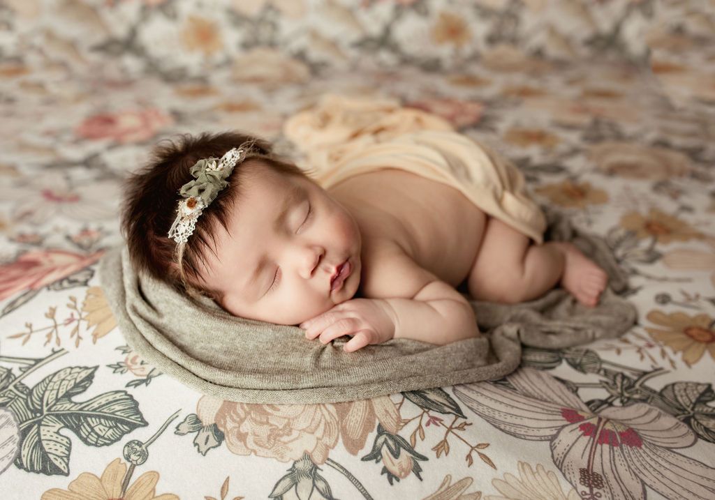 Newborn baby sleeping on floral backdrop, wearing a headband, wrapped in fabric.