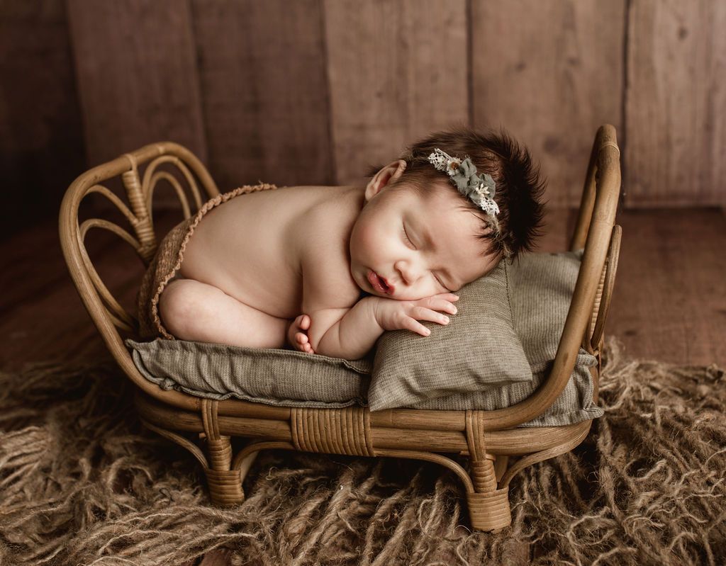 Baby asleep in a small woven bed, wearing a floral headband. Brown background, neutral tones.