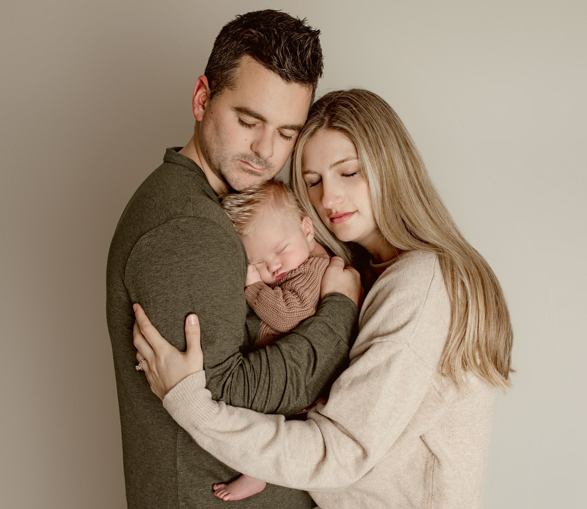 Parents embrace a sleeping newborn baby, eyes closed, against a plain background.