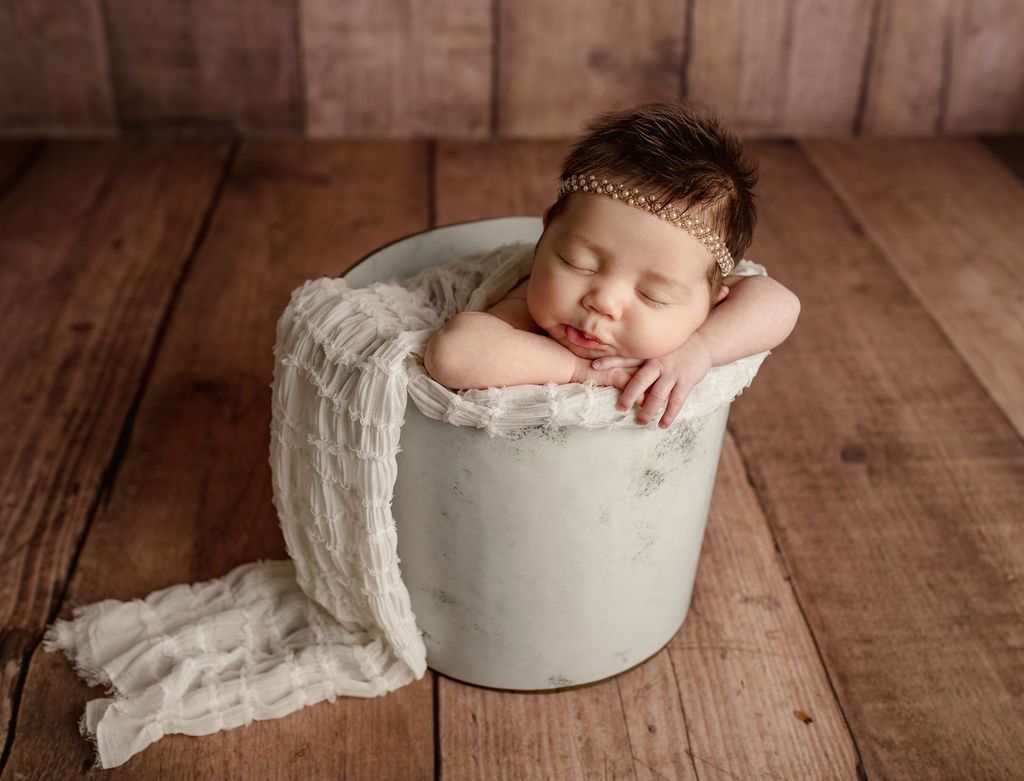 Newborn baby sleeping in a white bucket, wrapped in a white cloth, on a wooden surface.