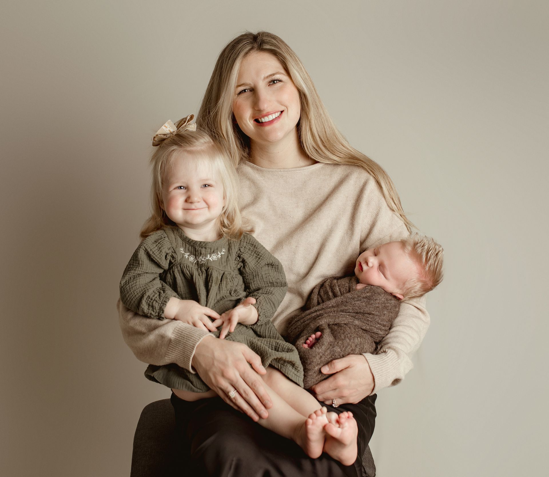 Woman smiles, holding a toddler and newborn baby. Indoor studio setting, light colors.