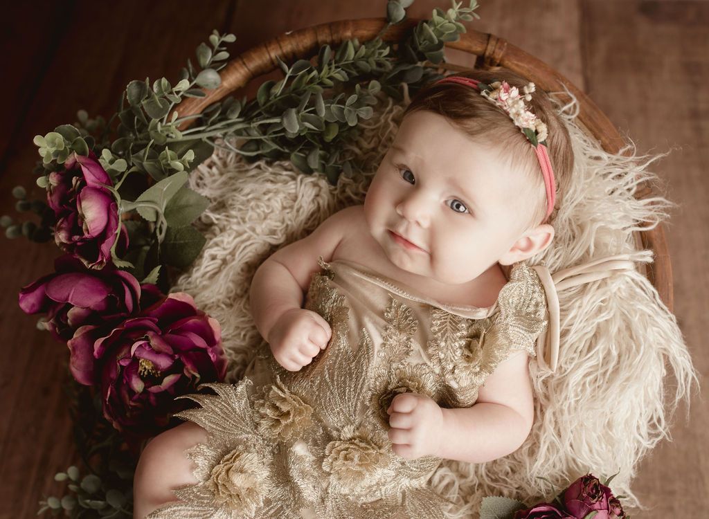Baby in gold dress, floral headband, and basket with flowers, looking up.
