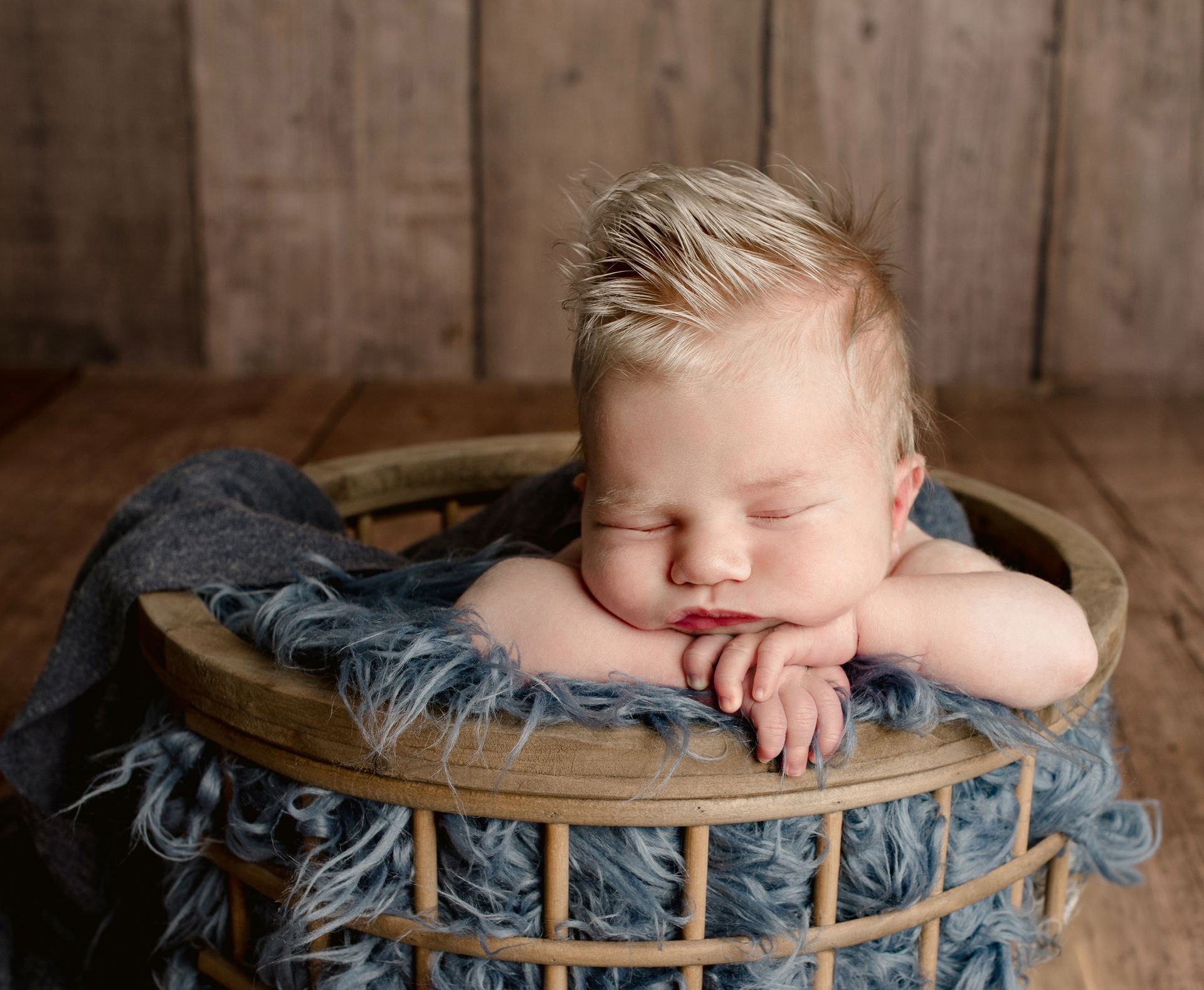 Baby with spiky blonde hair sleeping in a wooden basket lined with blue fabric.