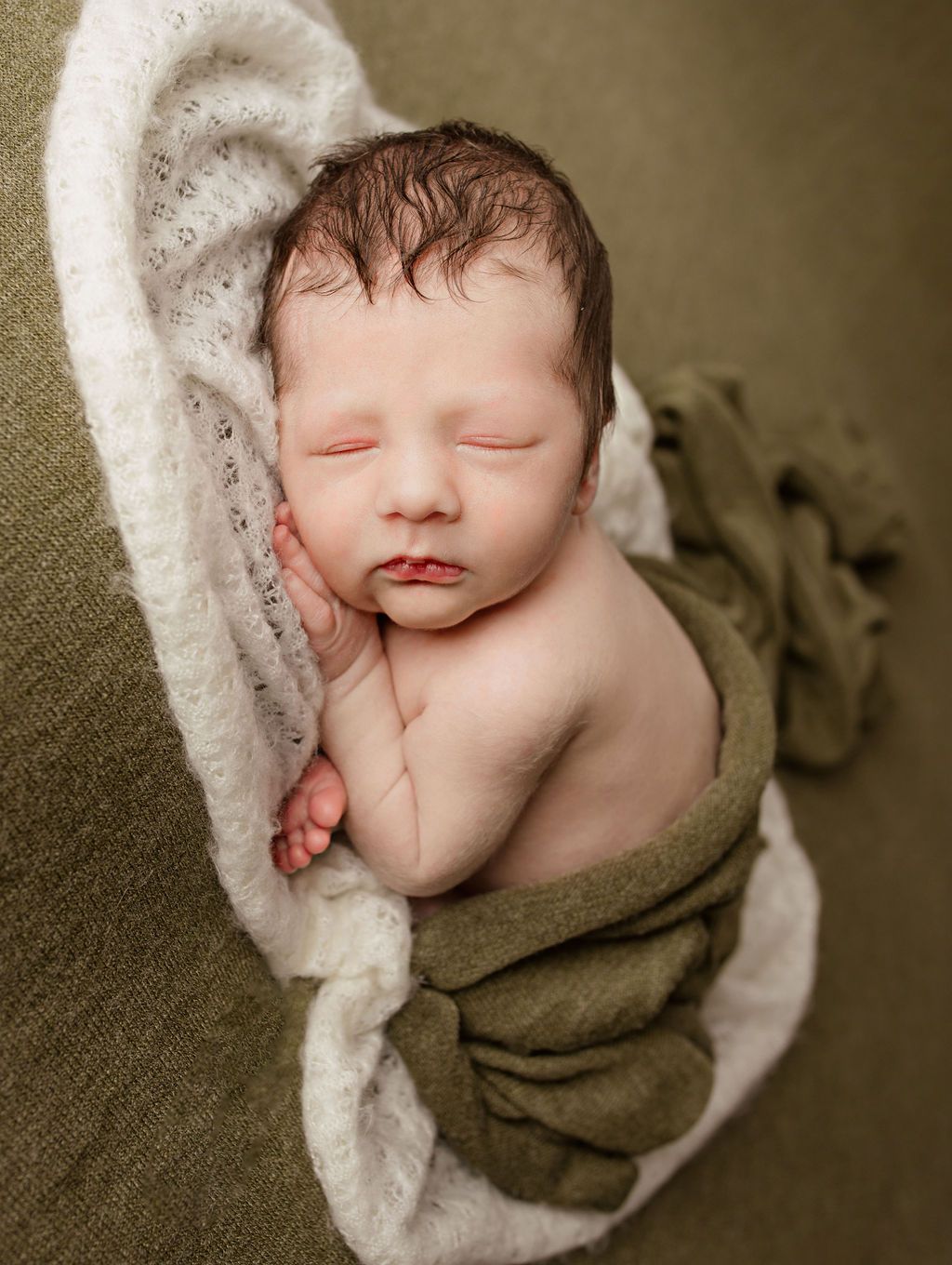 Newborn baby sleeping, swaddled in green and white blankets on olive-green fabric.