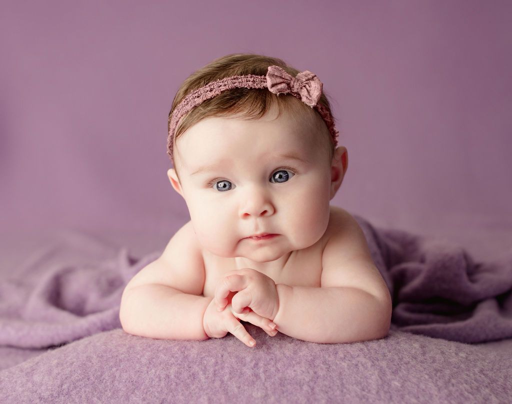 Baby with blue eyes, wearing a pink headband, resting on a purple blanket against a purple backdrop.