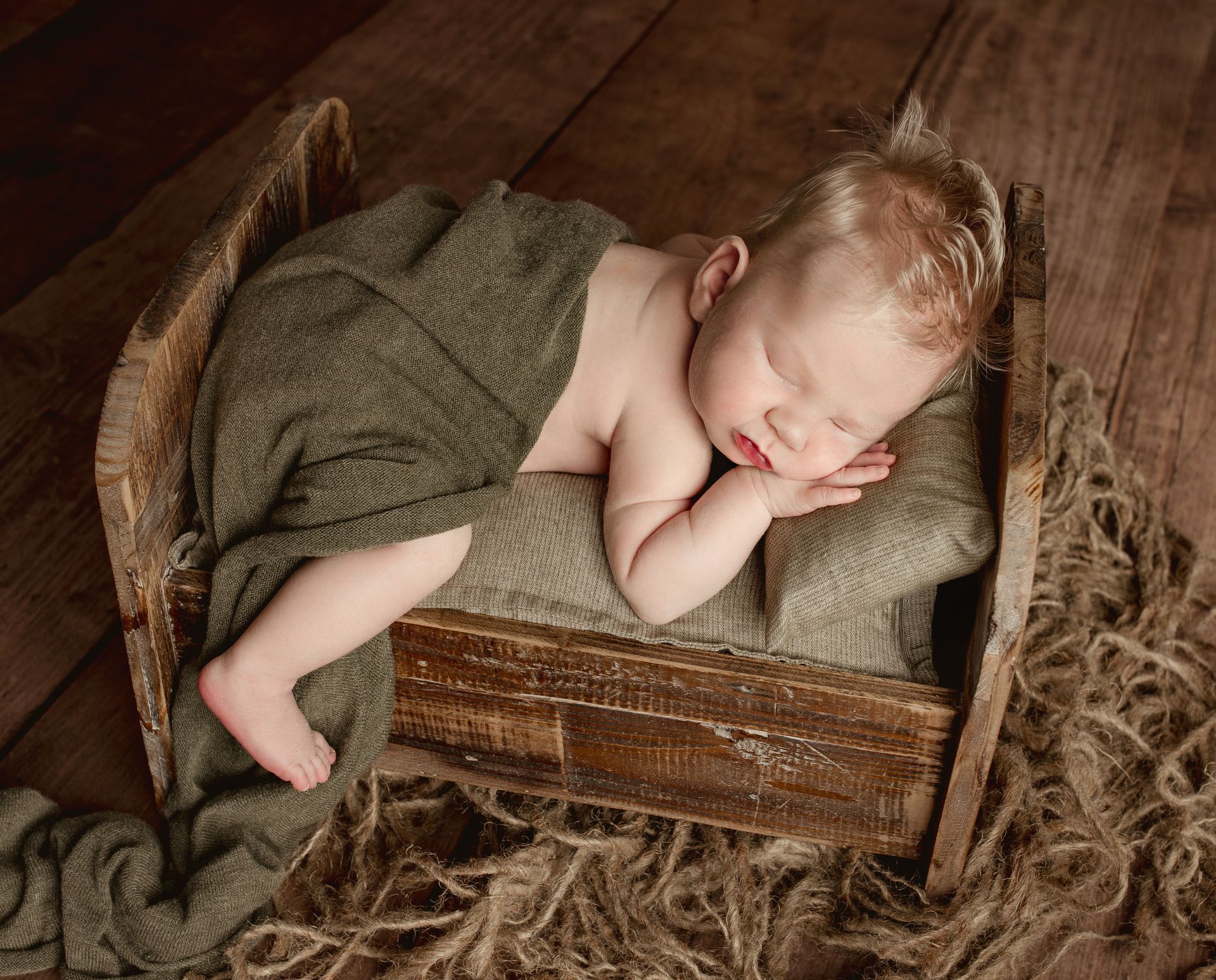 Newborn baby asleep in a rustic wooden bed, wrapped in a gray blanket.