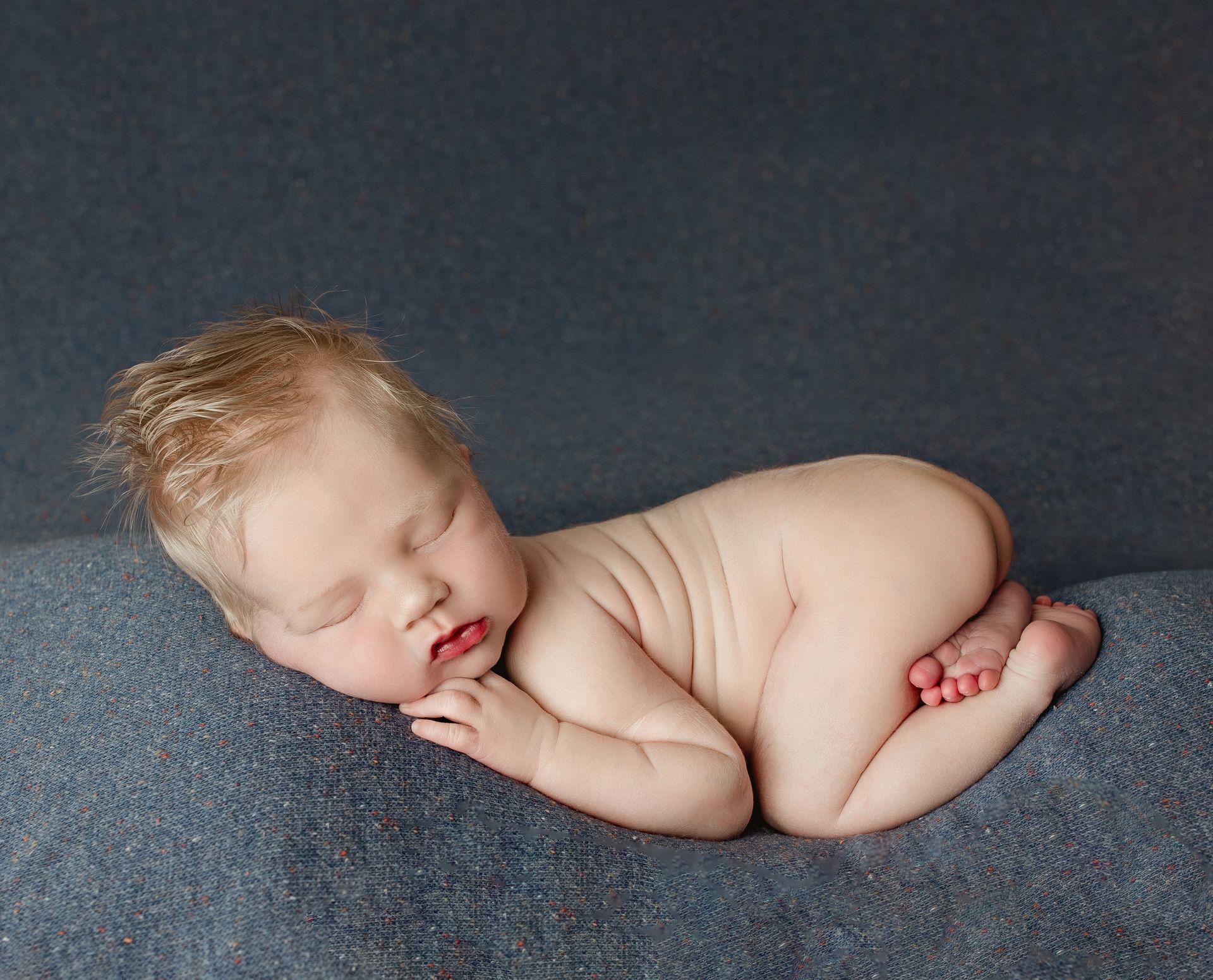 Naked newborn curled on a blue textured blanket, sleeping with closed eyes.