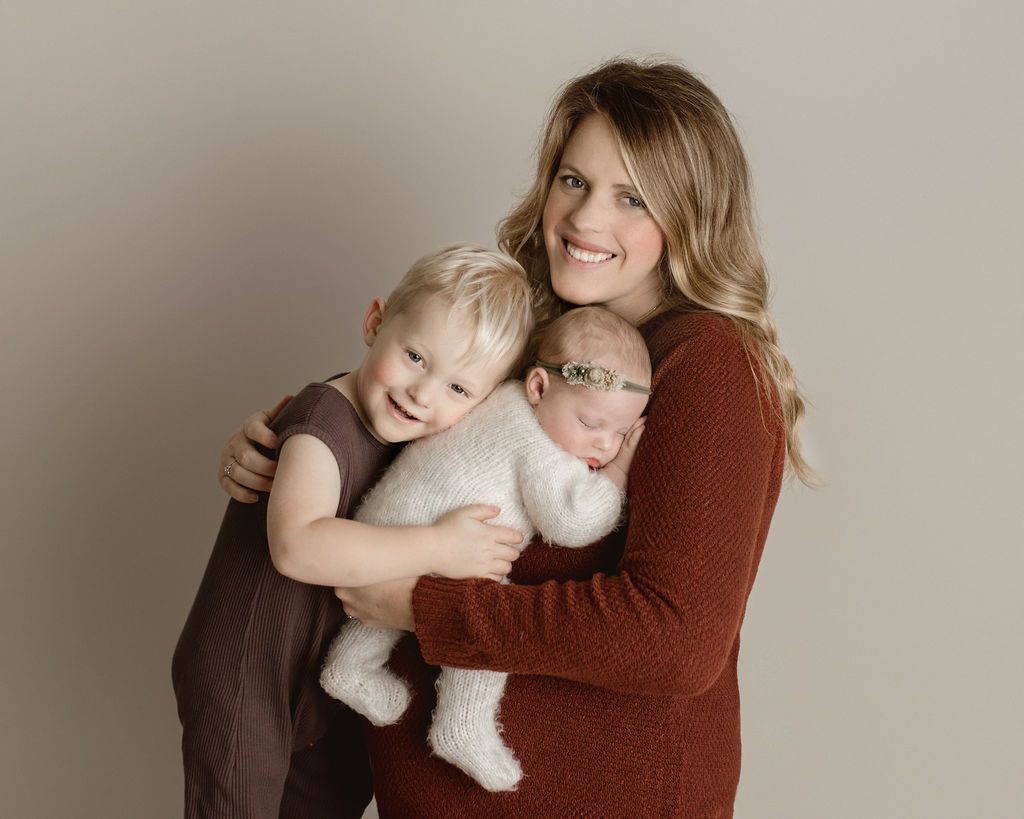 Mother hugs a baby and a toddler; all smiling. Neutral background. Baby in white, toddler in brown.