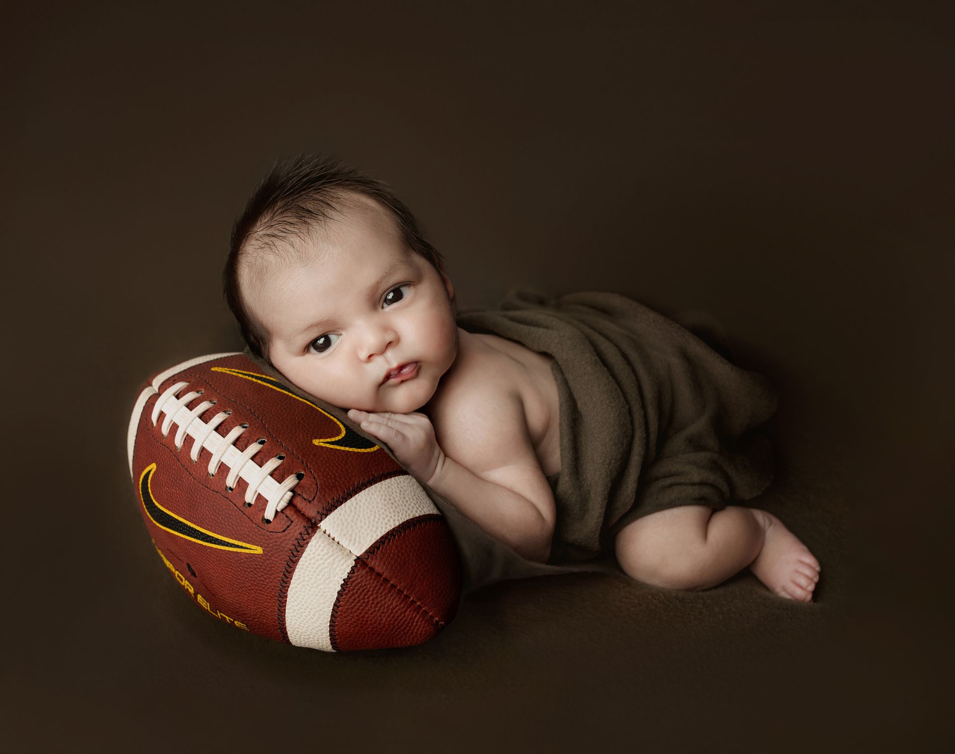 Newborn baby resting head on a football, wrapped in a brown blanket, brown background.