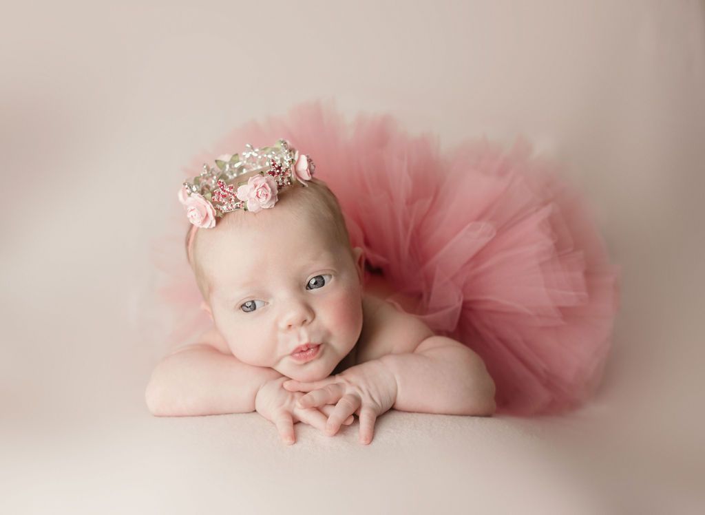 Baby in pink tutu and floral crown, resting on arms, light pink background.