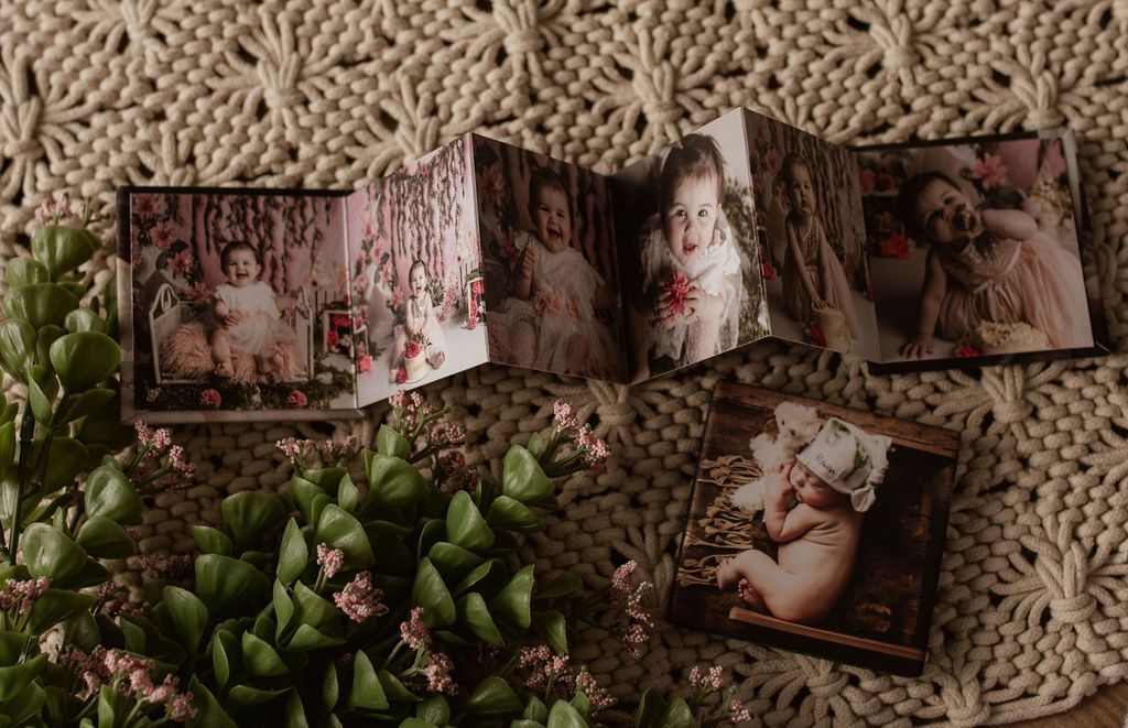 Accordion-style photo album with children's portraits, resting on a textured blanket, surrounded by greenery.
