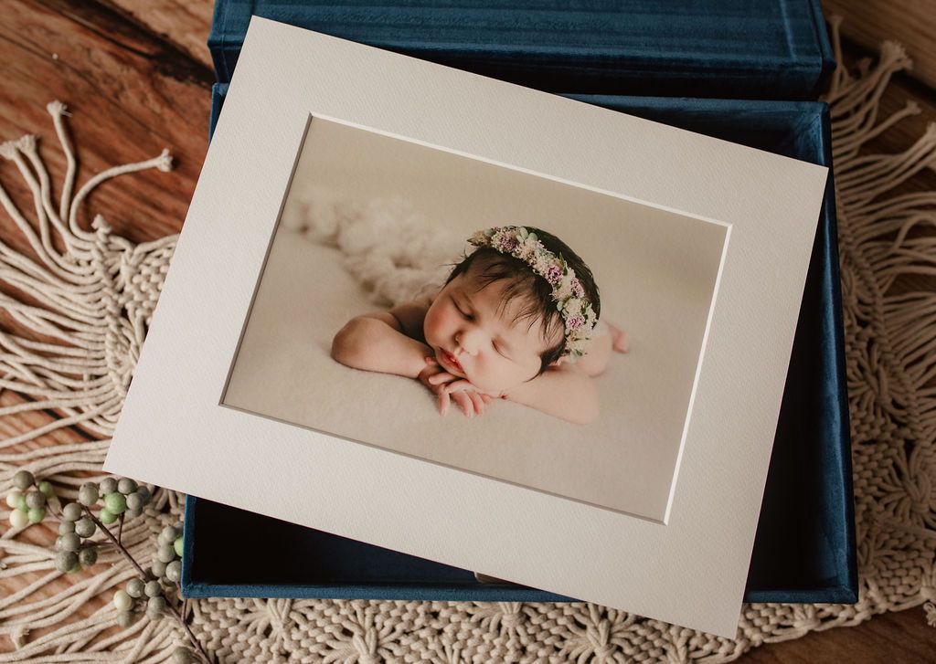 Newborn baby portrait framed in a mat, inside a blue box, on a wooden surface with decorative elements.