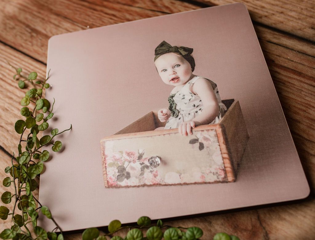 Baby in floral dress, sitting in a wooden drawer, with a bow on head. Light pink background.