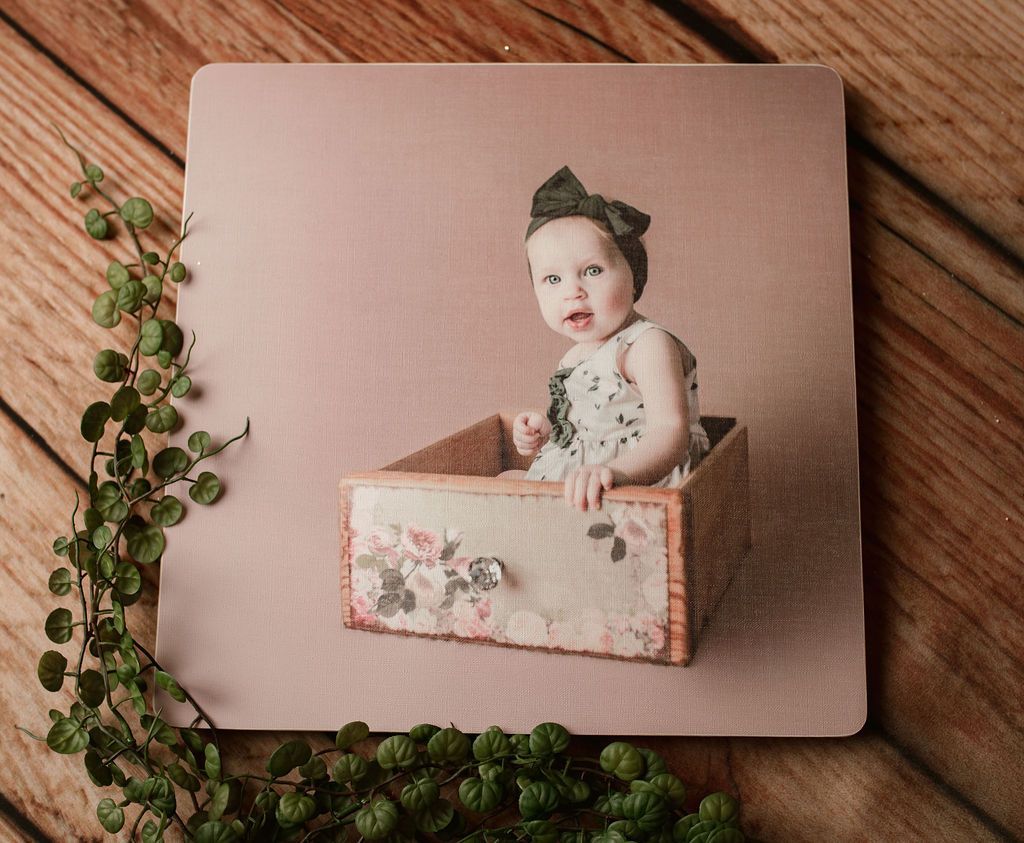 Baby wearing a bow in a floral dress sits in a decorative wooden box; pink backdrop.