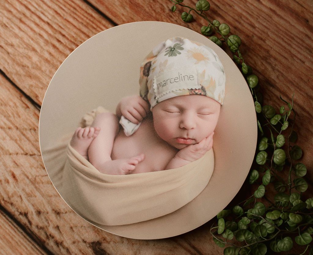Newborn baby sleeping, wrapped in beige fabric, wearing a patterned hat, on a wooden surface.