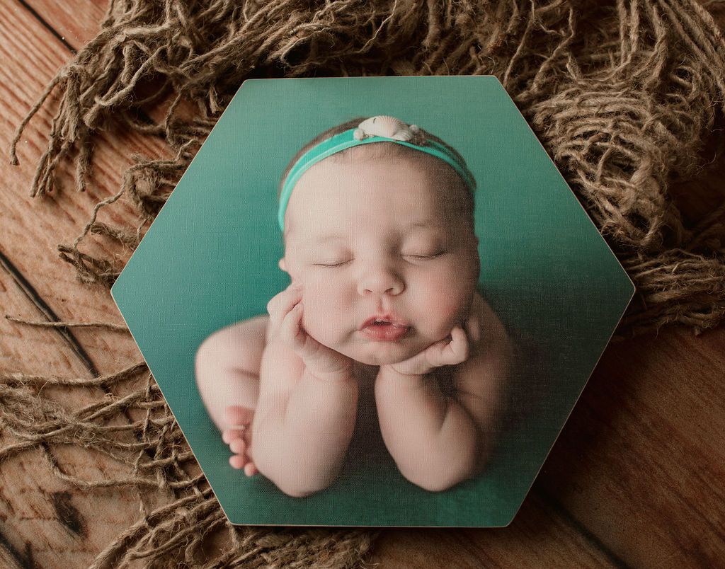 Baby with head resting on hands, wearing a teal headband, printed on a teal hexagonal tile, set on wood with rope.