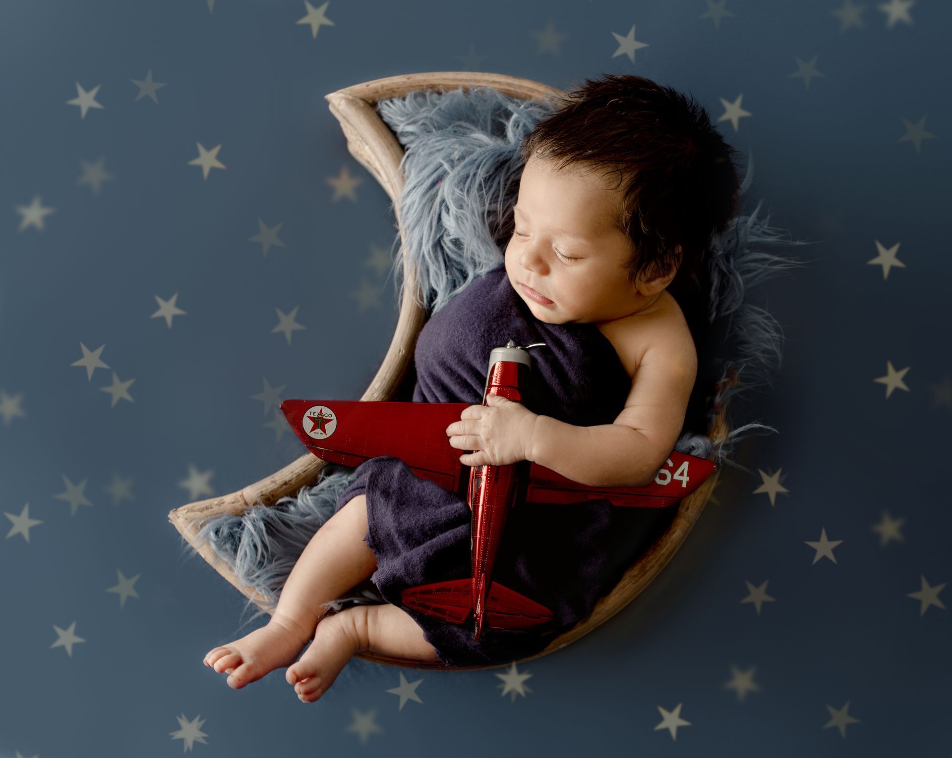 Baby sleeping in a crescent-shaped nest, holding a toy airplane. Blue blanket and star background.